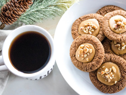 Plate of pecan gingerbread thumbring cookies next to a cup of coffee with pine cone decor in background