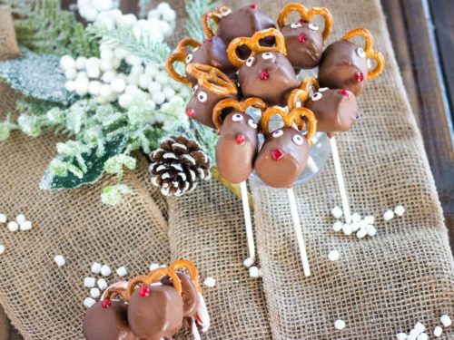 Huddle of chocolate reindeer marshmallow pops with mini marshmallow upright, with mini marshmallows and a few other marshmallow pops laying around them on burlap