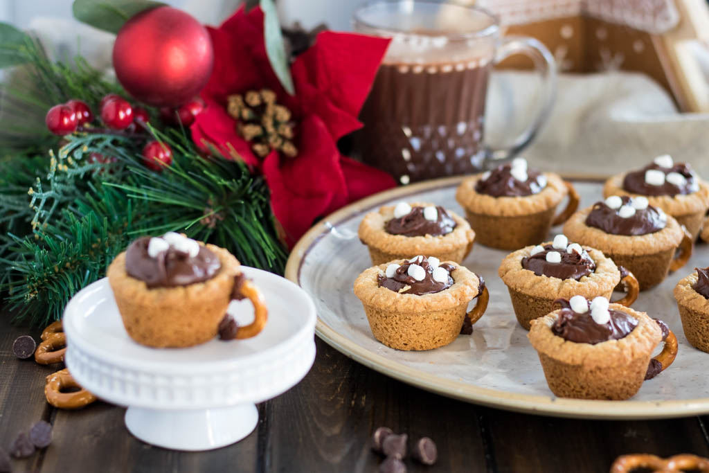 hot chocolate cookie cup on pedestal, next to plate of cookie cups, surrounded by chocolate chips, pretzels, poinsettia decor and cup of hot chocolate in background