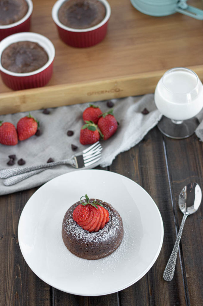 Un-cut lava cake with sliced strawberry on white plate with strawberries and chocolate chips, glass of milk, extra forks and more lava cakes in background
