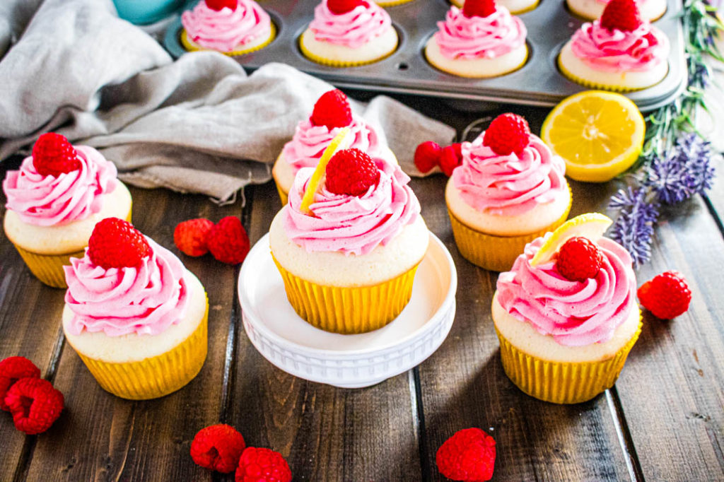 Single lemon cupcake with raspberry buttercream on white pedastal with cupckaes and raspberry on brown board and in muffin tin
