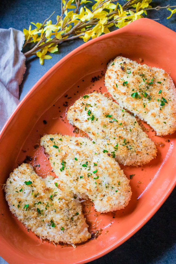 Garlic parmesan crispy baked chicken in orange oval baking dish with yellow flowers in background