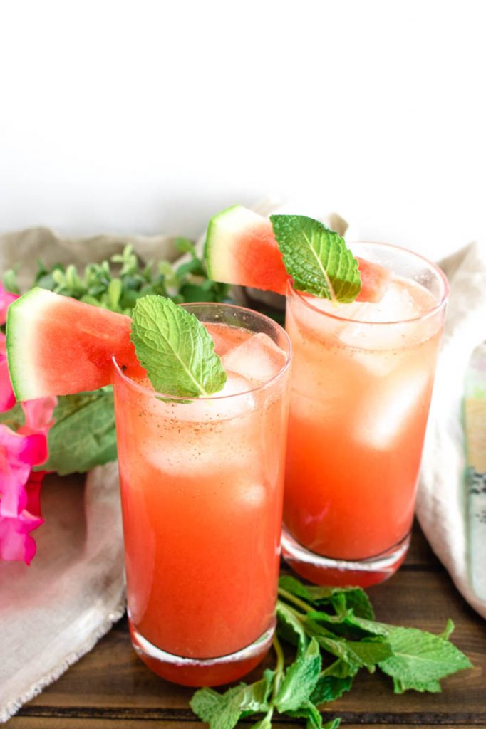 Two glasses of minty watermelon agua fresca with watermelon slice and mint garnish, on brown wood table. 