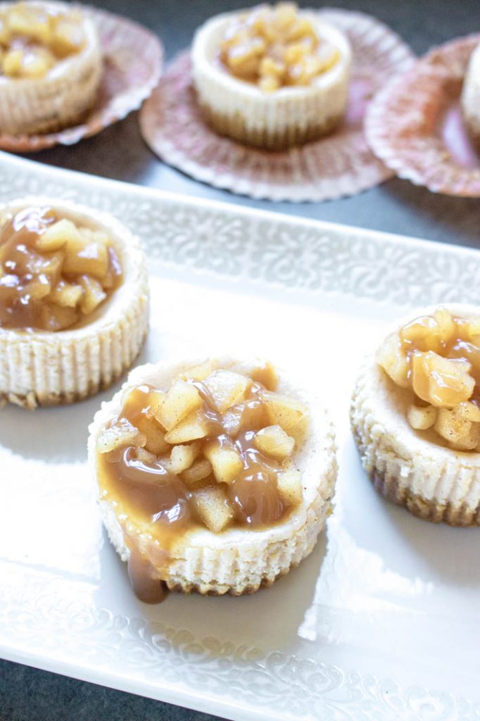 Spiced Apple Cider Mini Cheesecakes on white decorative plate, with three more behind in peeled down cupcake liners