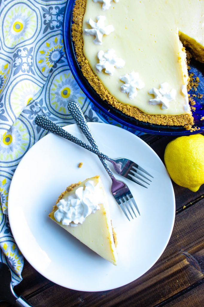 Slice of lemon icebox pie on round white plate with two forks. Full pie with one missing slice in top right corner with whole lemon just below. 