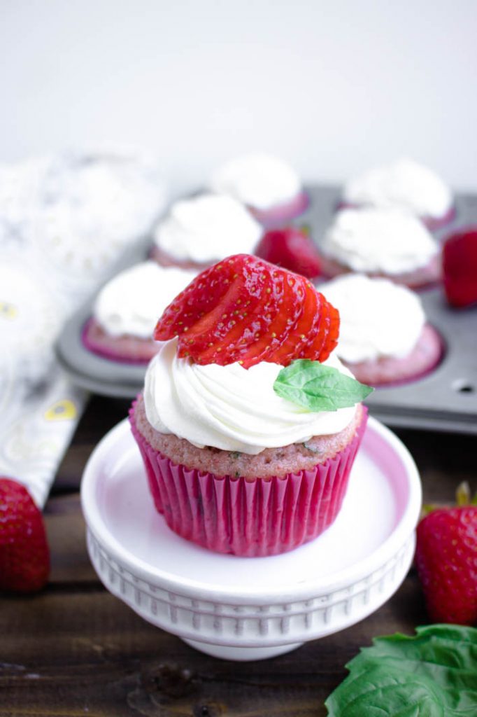 strawberry basil cupcake on white pedastal with frosted cupcakes in tin behind, and strawberries and basil at base 